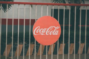 Okrągłe logo Coca-Cola zawieszone na metalowym ogrodzeniu przed budynkiem.