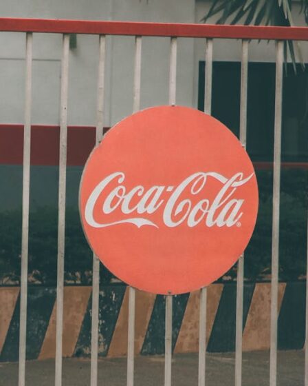 Okrągłe logo Coca-Cola zawieszone na metalowym ogrodzeniu przed budynkiem.