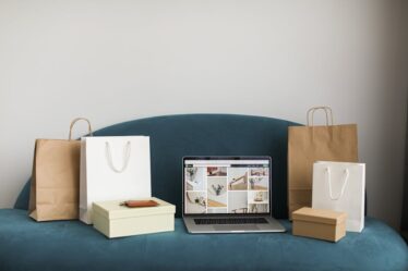 Laptop z otwartą stroną sklepu internetowego oraz torby i pudełka na kanapie.