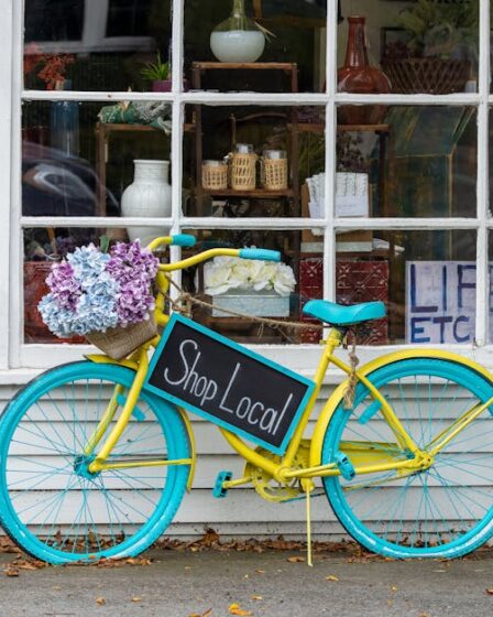 Żółty rower z turkusowymi kołami i tabliczką „Shop Local” przed witryną sklepową.