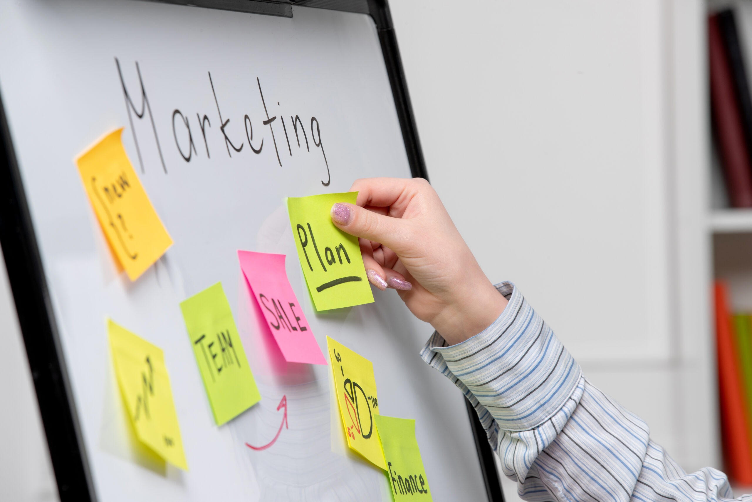 kobieca ręka przyklejająca sticky notes do białej tablicy z napisem "marketing"