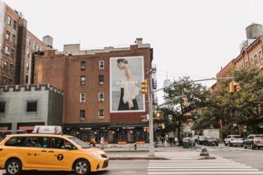 Żółta taksówka na skrzyżowaniu w Nowym Jorku z dużym billboardem Burberry na budynku.