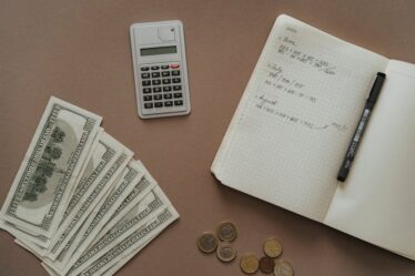 Kalkulator, notatnik z zapiskami finansowymi, banknoty oraz monety na brązowym tle.